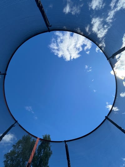 The image shows a view looking up through the netting of a trampoline towards a bright blue sky with scattered white clouds. The edge of the trampoline's protective netting forms a circular frame around the sky. Trees are visible in the lower left portion of the frame, seen through the netting. The scene appears to be outdoors on a sunny day. The location is identified as the city of Novozhilovo, Russia. There are no people or specific actions depicted in the image.