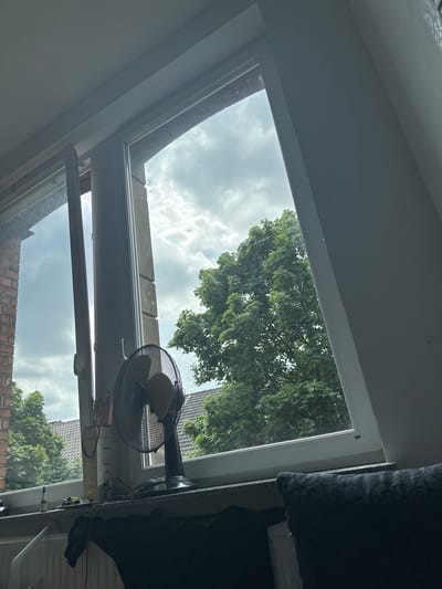 The image shows a close-up of a window with a fan on the windowsill. Outside the window, there are trees and houses. The sky is cloudy. The scene appears to be from a residential area in Hamm, Germany. There are no people visible in the image.