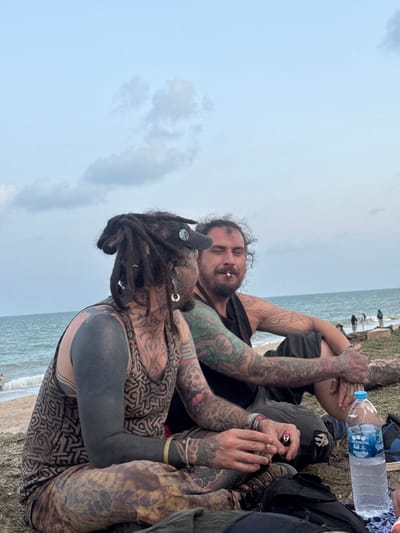 Two adult males are seated on dry ground adjacent to a sandy beach and the ocean in Ban Pang Pring, Thailand. The sky above is light blue with scattered white and grey clouds. The male on the left wears a brown and beige patterned sleeveless shirt, patterned pants, a dark cap covering dark dreadlocks, and multiple earrings. He displays extensive tattooing, including a solid black sleeve on his left arm and intricate patterns on his chest, neck, and right arm. He holds a small, dark cylindrical object in his right hand and faces slightly towards the second male. The male on the right wears a dark sleeveless shirt and dark pants. He has dark, curly hair, a beard, and extensive tattoos on both arms. He holds a lit cigarette in his mouth and looks towards the first male with a slight smile. A clear plastic water bottle with a blue cap rests on the ground to the right of the second male. The background features the ocean with small waves breaking on the sandy shore, where several blurred figures are visible in the distance.