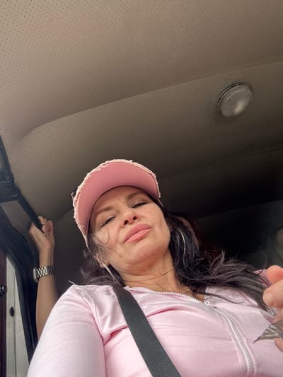 A woman in a pink baseball cap and pink zip-up jacket is in the passenger seat of a vehicle. She is looking down at the camera with her mouth slightly open. A hand is visible in the upper left corner holding onto a handle. The vehicle's interior, including the headliner and a light fixture, is visible. The scene is not identifiable as being in Каменномостское сельское поселение, Russia.