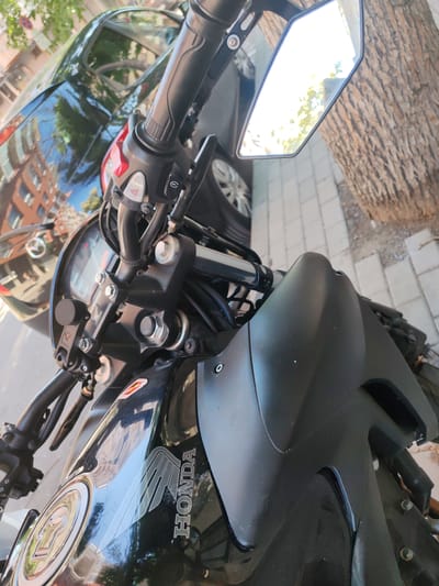 The image shows a close-up, angled view of a black Honda motorcycle parked on a sidewalk in Burgas, Bulgaria. The motorcycle is the primary subject, with details of its handlebars, mirrors, fuel tank, and engine visible. A black car is partially visible in the background, reflecting buildings and trees. A large tree with textured bark is to the right of the motorcycle. The ground is paved with light-colored rectangular tiles. The lighting suggests it is daytime, with sunlight reflecting off surfaces. No people are visible in the image.