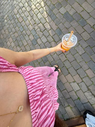 An overhead view depicts a person standing on a cobbled walkway, possibly in Hanau, Germany, though no specific landmarks are visible. The walkway is composed of small, dark gray cobblestones with patches of green moss or weeds growing in the cracks between the stones. The person is wearing a pink and white striped outfit and black sandals with gold accents. They hold a clear plastic cup filled with an orange beverage and a brown straw, decorated with panda motifs. A silver bracelet is visible on the person's wrist. A gold necklace with a pendant rests against the person's skin. The edge of a bench or raised platform is visible in the upper right corner of the image, along with a pile of folded clothes.