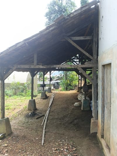 Under a rustic wooden structure, a dirt path stretches between two weathered buildings. The open-air shelter is supported by posts resting on concrete blocks, casting shadows on the ground. Bamboo poles lie along the path, leading towards foliage and glimpses of distant structures. A doorway is visible on the right building, while the scene suggests a rural or semi-rural setting.