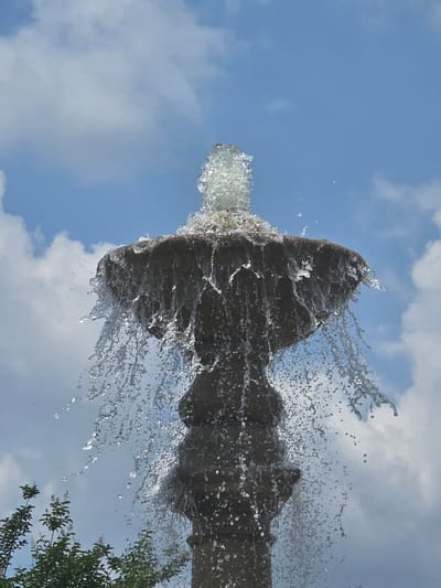 This is a low-angle, close-up, vertical shot of a water fountain against a partly cloudy blue sky. The fountain appears to be made of stone and is spraying water outwards and downwards, creating a dynamic cascade. The water is captured mid-splash, with numerous droplets and streams visible, catching the light. In the lower left corner of the image, the tops of green leafy trees are visible, suggesting an outdoor park or garden setting. The image is devoid of any people or animals. The scene appears to be located in Peachtree Corners, United States of America.