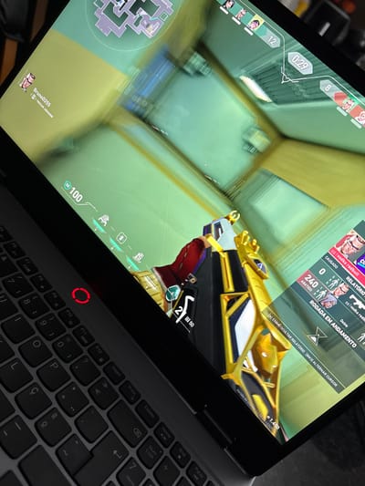 The image captures a close-up, angled view of a laptop screen displaying a first-person shooter video game, likely Valorant, with a partial view of the laptop's keyboard. The game is shown from a player's perspective, looking down a corridor or room corner with light green walls and a yellowish floor/ceiling. The player is holding a distinctive gold, black, and white weapon, which appears to be the "Prime Vandal" skin from Valorant.

Numerous in-game user interface (UI) elements are visible on the screen:
*   In the top left, a timer displays "0:29" and a number "8," possibly indicating remaining time in the round and players alive or rounds won.
*   Below the timer, player icons are seen, and a minimap is visible in the bottom left corner.
*   At the bottom center, the player's health is "100" and armor/shield is "50." Below this, icons representing player abilities are displayed.
*   The weapon's ammunition count shows "2" rounds in the magazine and "150" in reserve.
*   In the top right, a scoreboard or kill feed shows player names and scores, including "ggi vodka redbull," "CAUSADO 0," "ABATIDO 9," and "Debie." It also indicates "RODADA EM ANDAMENTO" (Round in Progress).
*   A message "[N] EXIBIR/OCULTAR RELATÓRIO [BM3] ALTERNAR CURSOR" (Show/Hide Report, Toggle Cursor) is also present.
*   The player's in-game currency or credits are "1.450."
*   In the very bottom left, the player's username "BrunoD255" is displayed along with "TROCAR JOGADOR" (Change Player).
*   The game version "Versão do c120242269" is visible in the bottom right corner of the screen.

The laptop's keyboard is partially visible in the bottom right of the frame, featuring a QWERTY layout with keys like F5 through F12, Insert, PrtSc, Delete, Home, End, PgUp, Num Lock, and several letter and symbol keys, indicating a Portuguese-Brazilian layout (e.g., 'Ç' key). A prominent red, circular illuminated indicator is present above the F10 key, which might be a power indicator or a special function key. The overall lighting suggests an indoor setting, with the screen being a primary light source. The image is taken from an elevated angle, looking down onto the laptop.