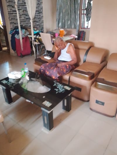 This image depicts an indoor residential living area in Mgbuoshimini, Nigeria. An elderly individual is seated on a light brown, two-seat sofa. The person, with short grey hair, wears a white sleeveless top and a patterned purple, orange, and black long skirt. Their left hand is raised to their face, covering the mouth and cheek area, while their eyes appear closed, suggesting rest or sleep. The right hand rests in their lap.

In front of the sofa is a black glass-top coffee table with dark legs and silver accents, featuring a circular opaque white insert and floral patterns. On the table are a clear plastic water bottle, a green plastic bottle, and a white tissue box or package. The floor is tiled with light-colored squares.

In the background, to the left, a wall with grey patterned wallpaper is visible, from which a white mosquito net or curtain hangs. Below this, a red suitcase topped with a pile of clothing is present. Further back, a light-colored table holds an orange thermos flask and a pink cup. A patterned blue and white curtain covers a window on the right side of the background.