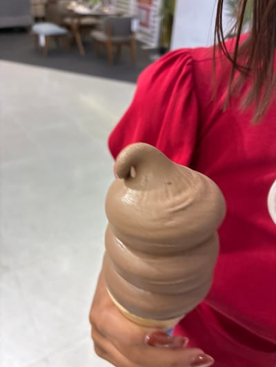 The image shows a close-up of a person holding a swirl of chocolate soft-serve ice cream in a cone. The person is wearing a red shirt, and their fingernails are painted a reddish-brown color. The ice cream is a light brown color and has a smooth, creamy texture with visible swirls. The background is blurred, but appears to be an indoor setting with some shelves and objects visible on the left. The lighting is bright, suggesting it could be daytime. The focus is entirely on the ice cream and the hand holding it. The image does not contain any visible text. The location context provided is Sao Thong Hin Town Municipality, Thailand.