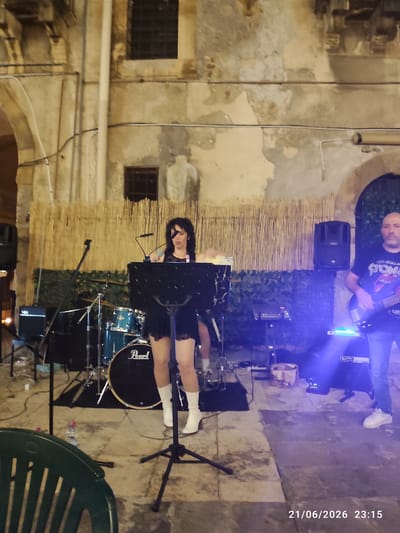In Militello in Val di Catania, Italy, a female singer stands at a music stand during an outdoor performance at night. She wears a black lace-trimmed dress and white boots. Behind her, a drum set with a "Pearl" logo is visible, along with bamboo fencing and a building facade. To her right, a man plays a bass guitar while wearing a t-shirt with a Rolling Stones logo and jeans. The scene is illuminated by stage lighting, casting blue hues. A green plastic chair is in the foreground, and a date and time stamp "21/06/2026 23:15" is present in the lower right corner.