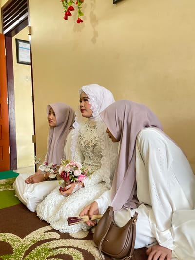 In the city of Pasar Paloh, Indonesia, three individuals are seated on the floor in a room. A woman in a white wedding dress and veil is positioned in the center, holding a bouquet of pink and red flowers. To her left, a woman in a light purple hijab looks away from the camera. To the right of the bride, another woman in a light purple hijab is partially visible, leaning forward and holding a fan. A brown handbag rests on the floor in front of her. The room has pale yellow walls, a wooden door on the left, and a patterned rug on the floor. Red artificial flowers are visible hanging from the wall in the upper left corner.
