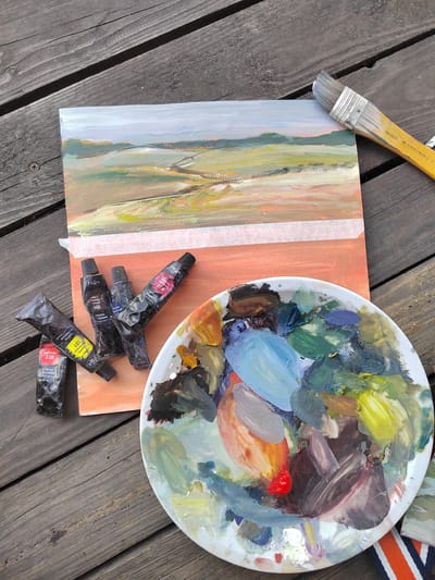 An overhead, close-up shot shows a painting of a landscape on a wooden table, near the village of Lynovitsy, in Belarus. The painting is on a piece of paper, taped to an orange surface, and is rendered in impressionistic strokes of green, brown, white, and pink. In front of the painting, a white, round palette is covered in dabs of oil paint in various colors, including blue, green, yellow, brown, and red. Several tubes of oil paint are arranged to the left of the palette and painting, with labels in Cyrillic script. One tube is red, labeled "218 Краска"; another is yellow, labeled "101"; and two others are black, labeled "606" and with blue text. A paintbrush with a yellow handle and grey bristles lies to the right of the painting. The wooden table surface appears weathered with distinct grain lines and visible knots.