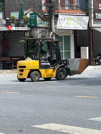 A yellow Komatsu forklift is parked on a paved road in what appears to be a commercial district in Tân Quang Commune, Vietnam. The forklift is positioned in the foreground, with its front facing left. A metal bin is attached to the lifting mechanism of the forklift, angled downwards. The forklift has a visible "KOMATSU" logo and the number "25" on its side. A conical Vietnamese hat (nón lá) is visible on top of the forklift. In the background, buildings with various signs line the street, including a green sign with white lettering that says "COFFEE PIZZA HERE" pointing left, and a large white banner with Vietnamese text and graphics, possibly advertising a nail salon. Power lines and electrical cables are strung across the top of the image. The road surface is asphalt, marked with yellow lines. Scattered on the road are fallen leaves. Other elements in the background include outdoor seating at what might be a cafe or restaurant, parked motorcycles, and architectural details of the buildings.
