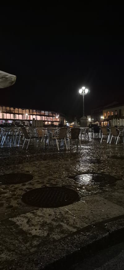 The image shows an outdoor cafe scene at night in the Plaza de Segovia, Navalcarnero, Spain. The cobblestone ground is wet, reflecting the lights from the surrounding buildings and a lamppost in the center of the plaza. Numerous tables and chairs, made of metal and wicker, are arranged in rows, many of them empty. In the background, a long, multi-story building with arcades and balconies is illuminated from within, suggesting shops or residences. A few indistinct figures can be seen through the windows of the building. The overall atmosphere is quiet and deserted, likely due to the late hour and wet conditions.