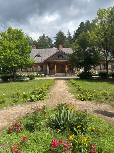 The image depicts a long, narrow, gravel pathway leading towards a rustic, wooden house. The pathway is flanked by lush green grass and flowerbeds bursting with red and yellow blossoms. Tall trees and dense forest form the backdrop to the house, with a grey, cloudy sky overhead. The house itself is a two-story structure with a wooden shingle roof, several windows with shutters, and a prominent chimney. A covered porch with a railing extends across the front of the house. Two wooden benches are visible on the lawn, one on the left and one on the right, inviting rest. The scene appears to be set in a rural or historical location, possibly a museum or heritage site, given the well-maintained pathway and manicured gardens leading up to the traditional architecture. The overall impression is one of tranquility and natural beauty, with a hint of rustic charm. The location is identified as Sielachowskie, Poland.