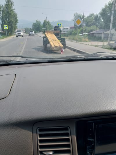 An overcast, daytime, outdoor, eye-level, medium shot captured from inside a vehicle shows a road and surrounding environment in the city of Belokurikha, Russia. The focus is on a trailer with wooden planks being towed by a tractor, moving away from the camera. On the left side of the road, a yellow diamond-shaped pedestrian crossing sign is visible, along with two cars. A second yellow diamond-shaped sign is visible on the right side of the road. Trees and a few buildings line the road. The foreground is dominated by the dashboard of the vehicle.