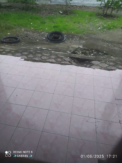 The image shows a tiled patio in Kebumen, Indonesia, with a crack running through the middle of the patio. The tiled patio is in front of a grassy area with two tires lying on the ground. There are no people in the image. The image was taken from a lower angle. The timestamp on the bottom right corner of the image says "01/06/2025 17:21." The image was taken on June 1, 2025 at 5:21 PM.