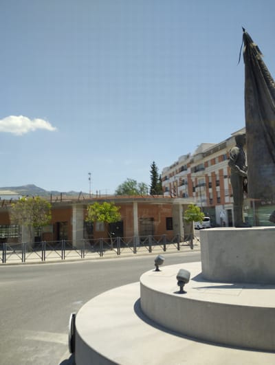 The image depicts a statue of a man in military uniform holding a flag in Cabra, Spain. The statue is situated on a circular concrete pedestal with two dark-colored lights embedded on its surface. The statue itself is made of bronze and is partially obscured by the flag. In the background, a multi-story building with orange and white facades can be seen, along with a single-story building with an orange exterior. The sky is clear and blue with a few white clouds. A road runs in front of the buildings and statue, and a metal fence separates the sidewalk from the street. There is also a small tree with green foliage planted on the sidewalk. The overall scene suggests a public space or monument.