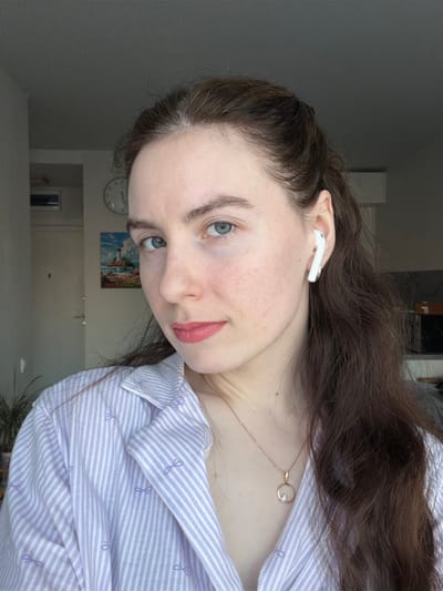 The image displays a headshot and upper torso of a female individual in an indoor residential setting within the city of Ковалёво, Russia. The subject, possessing fair skin, long dark brown hair, and light blue/green eyes, gazes directly at the camera with a neutral expression. She wears a light purple and white striped collared shirt featuring small bow patterns and a rose gold-colored necklace with a circular pendant containing a clear gemstone. A white wireless earbud is present in her right ear. The background shows a light-colored wall with a partially visible door on the left, an overhead rectangular vent, a circular wall clock, and a framed picture depicting a coastal scene with a lighthouse. A portion of a green potted plant is visible in the lower left foreground. The background to the right is darker and less defined.