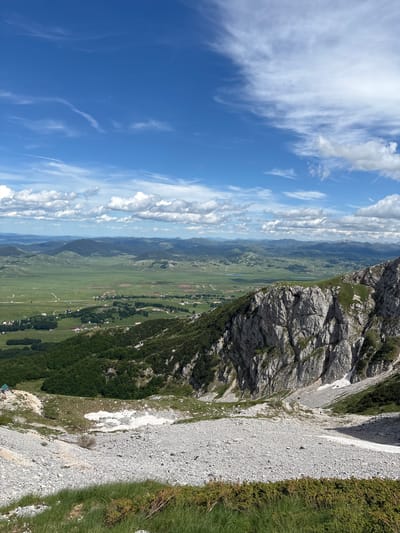 The image depicts a vast, mountainous landscape under a bright blue sky dotted with white clouds. In the foreground, a steep, rocky slope covered in loose grey stones and sparse green grass descends. Further down, a lush green valley stretches out, dotted with small villages and fields. The valley is bordered by rolling, tree-covered hills and distant, hazy mountains. The scene appears to be taken from a high vantage point, offering a panoramic view of the natural beauty of the area, likely in the vicinity of Pašina Voda, Montenegro.