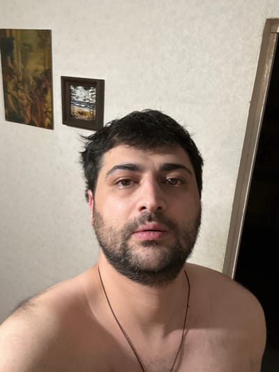 The media file is an image depicting a close-up, sideways portrait of a man with dark hair and a beard. He is shirtless and appears to be looking directly at the camera with a neutral expression. Behind him, the wall is a light, textured color, and two framed pictures are visible. The top picture is small and dark-framed, with a predominantly black and white image inside. Below it is a larger landscape painting with warm, earthy tones. A thin black cord or necklace is visible across the man's chest and neck. The lighting suggests an indoor setting, possibly at night or in a dimly lit room. There is no visible text in the image. The scene is set in Gyumri-Akhuryan, Armenia.
