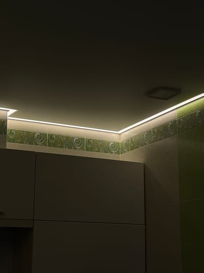 An interior scene captured in a low-light environment. The dominant features are dark brown kitchen cabinets situated below a tiled backsplash. The backsplash is composed of two distinct tile types: a lighter colored tile in the lower section and a decorative green tile with swirling patterns in the upper section. A prominent element is the strip of LED lighting installed along the ceiling-to-wall transition, casting a warm glow over the upper part of the cabinets and the backsplash. The ceiling is dark and features a square, recessed light fixture. The image provides no information about Linyovo, Russia.