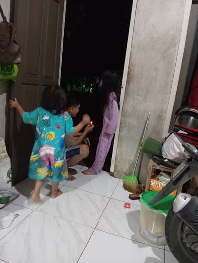 The scene depicts an interior space, likely a residence in Bulukandang, Indonesia, at night, viewed from the inside looking towards an open doorway. Three children are present. A young child, barefoot and wearing a multi-colored patterned nightgown, stands in the left foreground with their right hand near a brown wooden door frame. In the center, a second child, barefoot and wearing a light-colored top and dark shorts, crouches. This child holds a lit lighter or small flame between both hands, facing towards the right. A third child, barefoot and wearing light purple patterned pajamas, stands upright leaning against a light-colored door frame/wall on the right, observing the flame.
The indoor area features a white tiled floor with grey grout lines. To the left, a brown handbag with a "MK" logo and a green plastic bag hang on a wall, with a white sack resting on the floor below. To the right, a green broom leans against the wall, next to a small wooden stand holding various items. Partially visible in the extreme right foreground is the front section of a black and silver scooter or motorcycle. A small red toy car and a clear plastic bucket with a green lid are on the tiled floor. The outdoor area visible through the open doorway is dark, with faint distant lights suggesting a nighttime setting.