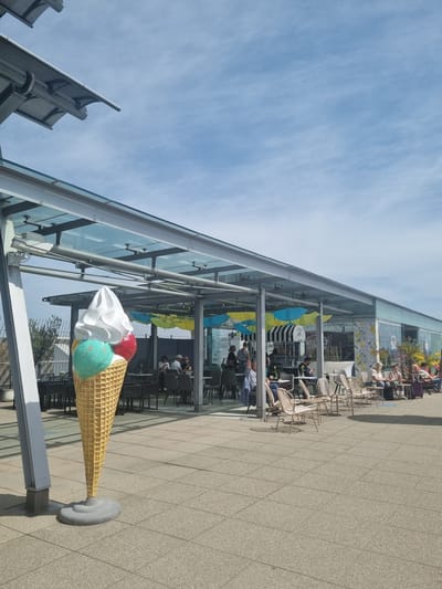 The image shows a life-sized decorative ice cream cone sculpture in front of a building in Echterdingen, Germany. The cone has a textured yellow waffle pattern and is topped with three scoops of ice cream: white, teal, and red. The sculpture is placed on a small gray stand. Behind the sculpture is a modern glass-walled building with tables and chairs inside, suggesting a restaurant or café. People are seated at some of the tables. Yellow and blue umbrellas are visible under the building's awning. The sky is blue with some clouds. The ground is paved with square tiles. Part of a structure with gray metal screens is visible on the left edge of the frame.