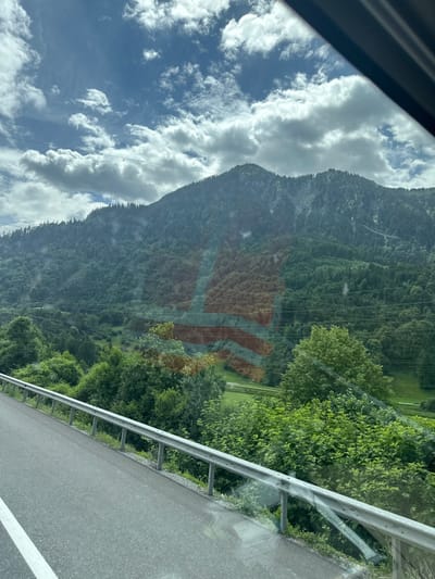 The image shows a view from a car driving along a road with a metal guardrail on the right side. The road is asphalt with a white line. On the left, a steep, heavily forested mountain slopes upwards, partially obscured by clouds. The sky is blue with scattered white clouds. The trees on the mountain are a deep green, and some power lines are visible running across the slope. In the distance, there appear to be some clearings or meadows on the mountainside. A subtle, large orange and red pattern, possibly a reflection or a sign on the mountain, is visible on the forested slope. The overall scene suggests a scenic drive through a mountainous region. The lighting indicates it is daytime, and the presence of clouds suggests variable weather. The location is Nenzing, Austria.
