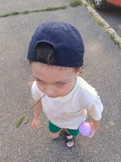 A young boy, wearing a navy blue baseball cap, a white t-shirt with a pocket, and green shorts, stands on an asphalt surface. He has a blade of grass in his mouth and is holding a purple spherical object in his right hand. The scene appears to be outdoors, likely in a residential area given the presence of a car tire and rim visible on the right side of the image, bordering a grassy patch and curb. The asphalt is textured with small pebbles and appears to be a pathway or sidewalk. The lighting suggests it is daytime, possibly late afternoon due to the angle of the shadows. There is no specific indication of the city of Kladno, Czechia, within the image itself.