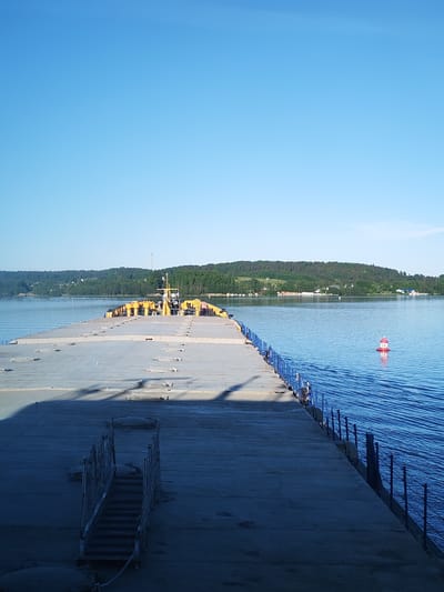 This image depicts a concrete pier extending into a calm body of water under a clear blue sky, located in Goritsy, Russia. The pier occupies the lower half of the frame, angled slightly to the right, creating a leading line towards the horizon. Its surface is light gray concrete, showing some wear and a distinct shadow extending from the left, indicating sunlight from the upper left. Numerous rectangular metal cleats or mooring points are embedded along its length. In the lower left foreground, a narrow set of dark metal stairs with ornate railings descends from the pier's edge.
Along the right side of the pier, a blue metal railing with vertical posts and horizontal bars runs its entire visible length. The water on the right is deep blue, exhibiting subtle ripples. Approximately mid-frame on the right, a red and white striped navigational buoy, cylindrical in shape with a rounded top, is visible, marked with the number "56".
Towards the far end of the pier, a section of yellow industrial equipment or a barge structure is docked or integrated. This yellow section features various mechanisms and what appears to be a control cabin or engine room. Beyond this, the water extends to a distant shoreline. The background consists of gently rolling hills densely covered with green trees and scattered patches of lighter foliage. A few small, distant buildings, including one prominent white structure, are visible among the trees on the far shore. The sky above is a uniform, bright light blue, with no clouds. No people are present in the scene.