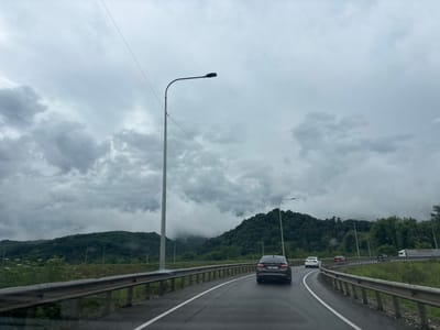 The image captures a highway scene under a cloudy sky, likely in or around ნიგოითის თემი, Georgia, given the context. The road curves to the right, bordered by a metal guardrail. Several vehicles are visible on the road, including a dark sedan in the foreground with a license plate that appears to be Georgian. Further ahead, a white car and a red car are also present, along with a white truck with "PIMAPEN" visible on its side. The landscape on either side of the highway features lush green hills and mountains, partially obscured by mist or low-hanging clouds. Tall streetlights line the side of the road, and a thin wire stretches across the sky, possibly from a power line. The overall atmosphere is overcast and somewhat subdued, suggesting a potentially rainy or humid day.
