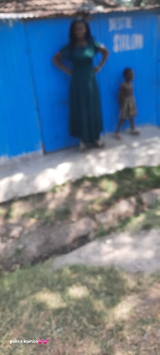 The scene, set in Homa Bay, Kenya, features two individuals positioned in front of a blue corrugated metal building. An adult female, centrally located, wears a dark green, short-sleeved, floor-length dress and stands with her hands on her hips. To her right stands a smaller child, dressed in light-colored clothing. The blue building's facade displays partially visible white lettering, indicating "BEST" and "SHALON" (or "SALON"). A corrugated metal roof is visible above the building. The ground immediately beneath the individuals is a light-colored, likely paved surface, which transitions into a foreground of unpaved earth, interspersed with sparse dry vegetation and a visible depression or trench. The image is blurred, captured under natural daylight conditions.