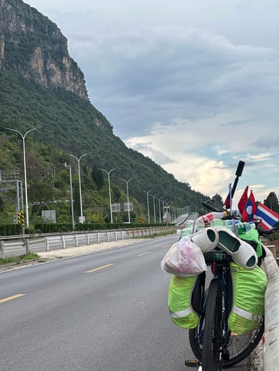 A bicycle loaded with luggage and flags is parked on the shoulder of a highway in Xishan District, China. The bicycle has two green panniers on the rear rack, a white plastic bag containing items, and a pair of white shoes secured on top. There are also several plastic bottles and other items attached to the bicycle. Two flags are visible on the rear rack: a red flag with a blue stripe and a white emblem, and a flag with red and white stripes. A black action camera is mounted on a stick above the handlebars. The highway stretches into the distance, bordered by a guardrail and a steep, tree-covered mountainside. The sky is overcast with scattered clouds. Streetlights are visible along the highway. The ground is paved asphalt with yellow lane markings.
