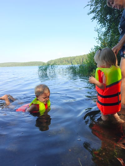 In Nepryakhinskoye Sel'skoye Poseleniye, Russia, two young children are in the water of a lake, wearing bright orange and yellow life jackets. One child is floating on their stomach, with their legs extended behind them, while the other child stands in shallow water, facing away from the camera. A woman, partially visible, stands behind the second child. The environment is a lake with a wooded shoreline under a clear blue sky.