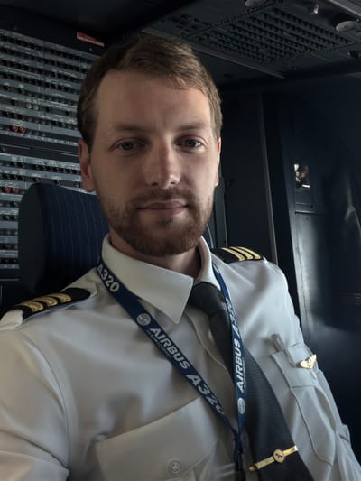The image depicts a male pilot in the cockpit of an Airbus A320 aircraft. He is wearing a white pilot's uniform with four gold stripes on his shoulders, a gray tie with a gold tie clip featuring a bird, and a blue lanyard with "AIRBUS A320 AN EADS COMPANY" printed on it. His uniform also has a gold winged pin on the left breast. The pilot has short brown hair and a beard.
The cockpit is visible in the background. To the left of the pilot is a control panel with numerous switches and buttons, including those for lighting, fuel, doors, and flight controls. The panel also displays a "CAUTION HIGH VOLTAGE INSIDE" warning. The seat is dark blue with pinstripes.
Based on the aircraft type and the logo on the lanyard, it can be inferred that the location is likely at an airport that handles Airbus A320 aircraft. It cannot be determined if this is Schiphol, Netherlands.