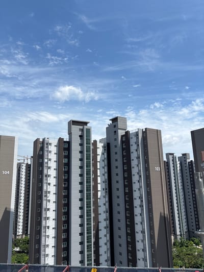 In Cheongju-si, South Korea, a series of tall, modern apartment buildings dominates the scene under a partly cloudy blue sky. The buildings are primarily colored in shades of gray and brown, with white accents and numerous windows. Some buildings display identifying numbers: '104' is visible on the leftmost building, and '103' is on a building further to the right. A construction crane can be seen behind the buildings on the left, suggesting ongoing development in the area. The perspective is from a higher vantage point, looking across the cityscape. Lush green trees are visible at the base of some buildings, adding a touch of nature to the urban environment. The overall impression is one of a densely populated residential area with contemporary architecture.