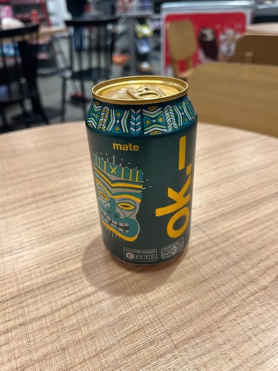 A dark teal or green beverage can is lying horizontally on a light-colored wooden table surface. The can has a shiny gold-colored top and a distinctive design featuring a geometric pattern in teal, yellow, and white around the rim. The main body of the can prominently displays large yellow letters spelling "ok-i". Below this text, also in yellow, is the word "mate". A stylized, yellow, grey, and teal tiki-like face graphic is depicted on the can.

On the right side of the can, a small white box contains text related to product grading. Visible text includes "ECO-SCORE" and an "A-BBBC" rating, along with an "ABCDE" scale where "A" and "B" appear to be highlighted.

The table surface shows a visible wood grain pattern. The background beyond the table is blurred, revealing indistinct shapes and colors that suggest an indoor environment, possibly shelves or other items in a room or store. The lighting appears to be artificial, casting a subtle shadow from the can onto the table. The scene takes place in Schweinfurt, Germany.