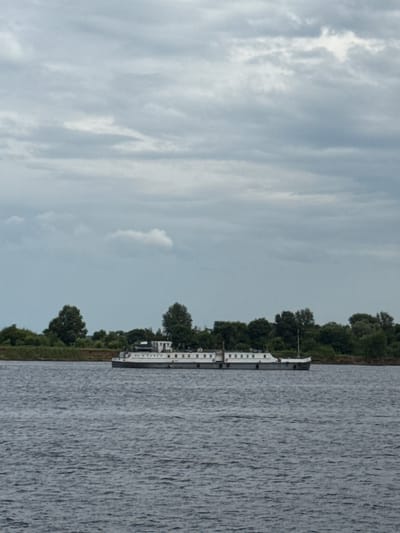 The image shows a wide river with a large, flat-topped white barge moving on it. The barge is elongated and appears to be a cargo vessel. It has a series of windows along its upper deck and a superstructure towards the stern. The river is calm, with ripples on the surface reflecting the overcast sky. In the background, a lush green treeline with scattered buildings is visible on the riverbank. The sky is filled with dense, gray clouds, suggesting an overcast or possibly stormy day. The overall scene appears to be a natural landscape with a functioning waterway. The location is identified as the city of Bet'ki, Russia.