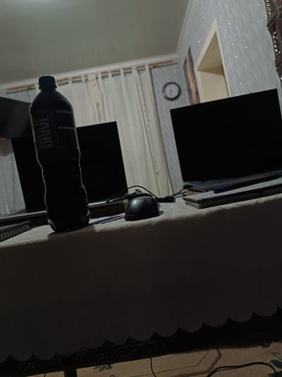 The image depicts a dimly lit indoor scene, likely a home office or study area. In the foreground, a dark bottle of what appears to be an energy drink, labeled "GAME DRIVE," stands prominently on a table covered with a patterned tablecloth. Next to the bottle, a computer mouse is visible. Two laptops are present in the background, their screens dark. A clock hangs on a wall adorned with sparkly wallpaper, and a doorway is partially visible. Curtains hang in the background, suggesting a window. The lighting is low, casting shadows across the objects and furniture. The overall atmosphere is that of a quiet, possibly late-night, workspace. The image does not provide any specific visual cues or context to place the scene within the city of Medveja, Moldova.