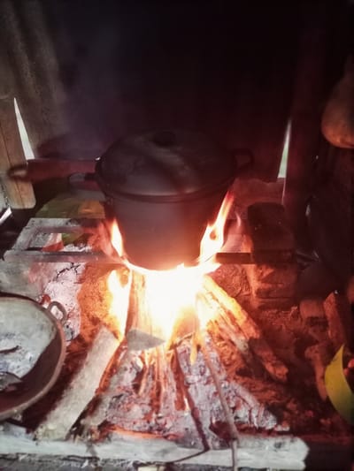 A black pot sits on a rustic stove made of bricks and wood, with a fire burning underneath. The flames are bright orange and yellow, licking up at the pot and casting a warm glow on the surroundings. The scene appears to be outdoors, possibly in a rural setting given the natural materials used for the stove. The time of day is unclear, but the dim lighting suggests it might be evening or a very overcast day. The pot is the main subject, positioned centrally, with the fire providing a dynamic element. The background is dark and indistinct, with some vertical elements that could be part of a shelter or structure. Another cooking vessel, a shallow, wide pan, is visible in the lower left corner, also appearing to be made of dark material. The overall impression is one of simple, traditional cooking. There is no visible text in the image. The setting is Gampong Teungoh, Indonesia.