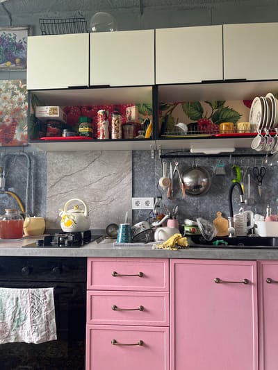 The image shows a cluttered kitchen in Khor, Russia. The kitchen features pink cabinets, white upper cabinets, and a gray countertop. On the countertop, there is a black gas stove with a white teapot, a jar of red liquid, and various kitchen utensils. The cabinets are filled with dishes, jars, and other kitchen items. A black oven with a towel hanging from it is visible on the left. The walls are covered with gray tiles, and there are decorative elements like paintings and wallpaper with floral patterns. The overall impression is of a lived-in and functional kitchen space. There are no people visible in the image.