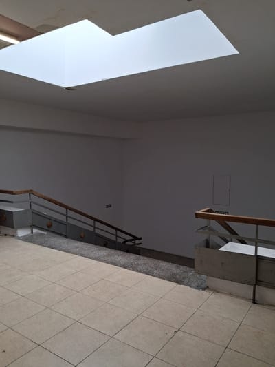 The image depicts an interior view of a stairwell, likely in a public building in Constanța, Romania. The perspective is looking upwards, emphasizing the architecture and the space.
The primary subjects are the staircase and its surroundings. The staircase features metal railings with a wooden handrail, supported by concrete structures. The treads of the stairs are visible, and the walls are painted a neutral color. A large, angular skylight dominates the left side of the image, flooding the upper portion of the stairwell with bright, natural light, suggesting it is daytime. The floor is tiled with large, square, beige tiles arranged in a diamond pattern.
There are no people visible in the image. The scene appears to be a quiet, empty interior space. Notable details include an electrical outlet and a small square access panel on the wall. There are also two small pictograms on the wall, one indicating an exit or path upwards, and the other an arrow pointing downwards, suggesting navigation within the building.
The overall impression is one of functional architecture, with a focus on light and space. The time of day is clearly daytime due to the strong illumination from the skylight. The weather outside is not discernible. The location context is Constanța, Romania. There is no discernible emotional tone conveyed by the image.