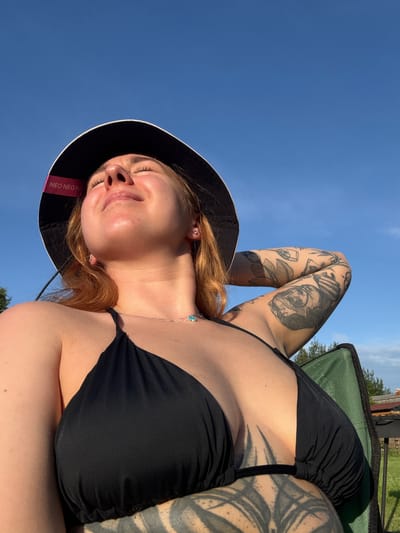 This is a close-up, low-angle shot of a young woman wearing a black bikini top and a black bucket hat with a pink band. She is lying down outdoors on a sunny day. The blue sky fills the background, with a hint of green foliage and some wooden structures visible at the top right, suggesting an outdoor recreational area or a backyard. The woman has her eyes closed and a relaxed smile on her face, indicating she is enjoying the sunshine. Her arms and chest are visible, and she has numerous tattoos on her arms and torso. The band on her hat has the text "NEO NEO NE" printed on it in white letters. The scene is illuminated by bright sunlight, casting shadows and highlighting the texture of her skin and tattoos.