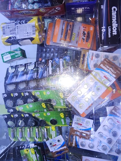 The image displays a close-up, angled view of various types of batteries, predominantly button and cylindrical cell batteries, packaged in blister packs and cardboard boxes. The items are densely arranged, with some overlap and glare from lighting. The batteries are of different brands, including "Camelion," "GoldenPower," and what appears to be "GP" and "Murata Silver Oxide." Packaging colors range from bright yellow and green to blue and black. Some of the larger cylindrical batteries are in yellow and black packaging. The overall impression is of a retail display or storage area for batteries. The scene is indoors, and the lighting is bright, causing significant reflections and glare on the plastic packaging. There are no people or recognizable landmarks visible. The location in Ushtobe, Kazakhstan, is not discernible from the visual information in the image itself.