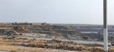 The image depicts a large-scale excavation site, likely a quarry or mine, located in the outskirts of Margilan City, Uzbekistan. The foreground is dominated by dry, sparse vegetation and piles of dirt and rocks. In the mid-ground, the excavation reveals layered geological strata, with steep, cut earth faces forming terraces. Several large, utilitarian buildings with light-colored roofs are visible in the background, situated on a level plateau above the excavation. These structures suggest industrial or storage facilities associated with the mining operation. The overall environment is arid and dusty, with a hazy sky overhead, indicative of a dry climate. The scene is devoid of any visible human activity or movement. A utility pole is present in the right foreground, partially obscuring the view of the excavation.