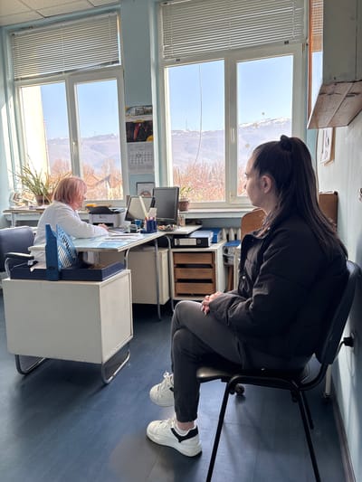 This media file depicts an indoor office setting, likely in Pirdop, Bulgaria, characterized by strong natural light streaming through large windows. The room features light blue walls, a dark blue floor, and light-colored office furniture.

Two women are present. In the foreground, a woman with long, dark hair, wearing a black puffer jacket, dark pants, and white sneakers with black accents, is seated on a tall, black office chair. She is leaning forward, looking down towards a desk area, with her hands clasped.

Further into the background, another woman with reddish-blonde hair and wearing a white coat (possibly a lab coat or professional attire) is seated at a white desk. She is actively working, engaged with a computer setup that includes monitors and a "Brother" brand printer. Various office supplies, such as a blue "PRIMA" binder and pen holders, are visible on her desk. A calendar featuring images of paragliders and the year "2026" is affixed to the wall, with visible dates including "1, 2, 4, 13 14 15 16 17 18 19, 20 21 22 23 24 25, 27 28 29 30."

The large windows offer a clear view of an outdoor landscape under a bright, sunny sky. The scenery includes bare-branched trees, suggesting a late autumn or winter season, and potentially distant hills or mountains. Decorative snowflake stickers are affixed to a window pane. Blinds are partially closed on some window sections. A potted plant adds a touch of greenery near a desk. The overall atmosphere suggests a focused, work-oriented environment during daylight hours.