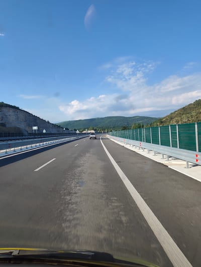 The image shows a highway in Peshtera, Bulgaria. The highway is a multi-lane road with clear white lane markings. There are guardrails on both sides of the road, and green sound barriers are installed along the right side. The landscape consists of rolling hills covered in trees, with a rocky embankment on the left side of the highway. The sky is blue with scattered white clouds. A white SUV is visible driving on the highway in the distance. The photo appears to be taken from inside a vehicle, with the dashboard visible in the foreground. The overall scene suggests a clear, sunny day on a modern highway infrastructure.