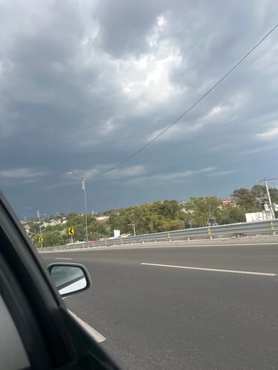 View from inside a moving vehicle on a highway in San Francisco Totimehuacán, Mexico. The road surface is dark gray asphalt with a white lane marker. A metal guardrail runs parallel to the road, interspersed with green trees and buildings. In the distance, utility poles are visible against a cloudy sky. The side mirror of the car is partially visible in the foreground.
