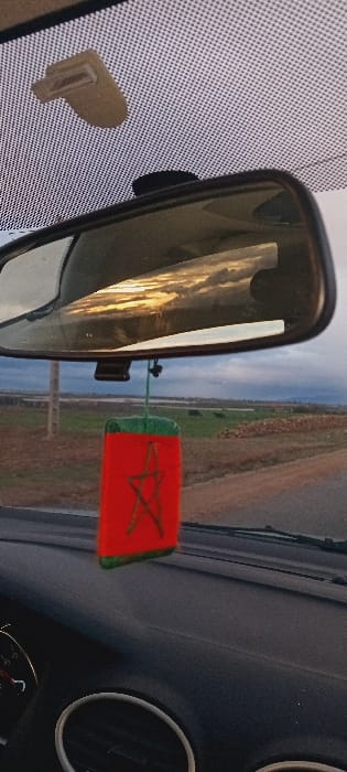 A car interior is shown. The rearview mirror reflects a landscape with water and sky. A red air freshener with a green outline and star design hangs from the mirror. Part of the dashboard is visible with circular vents. A beige object is attached to the windshield. The dashboard appears dark, and the reflection in the mirror shows an outdoor scene.