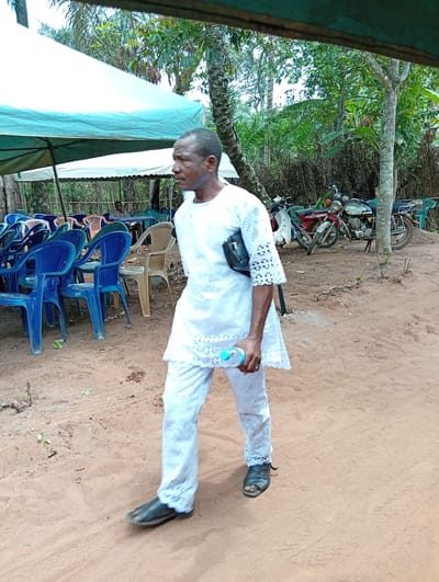 The image captures an outdoor scene, likely a temporary gathering or event in Umundugba, Nigeria. The ground is a light brown dirt or sand, showing some footprints and tire tracks. In the foreground, a man, appearing to be of African descent, walks from left to right. He is dressed in a clean, white, two-piece traditional or semi-traditional outfit, featuring intricate lace-like cutouts on the sleeves and the bottom edges of both his long top and trousers. He wears dark open-toed sandals and carries a small black crossbody bag over his left shoulder. In his right hand, he holds a clear plastic water bottle with a light blue cap. His gaze is directed slightly ahead and to his right, with a neutral expression.
The background features several elements indicating a communal setup. To the left, under a large canopy tent with a white top and light blue underskirt, numerous plastic chairs are arranged. The chairs are mostly blue, with some beige and orange ones, suggesting seating for attendees. Further back, beneath another tent structure, a person can be vaguely seen seated at a table. On the right side of the frame, behind the walking man, two motorcycles are parked on the dirt, one red and one with white and dark colors. Lush green foliage and various trees, including palm trees, form the backdrop, indicating a natural, possibly rural or semi-urban environment. The lighting suggests it is daytime. No visible text or prominent branding is discernible in the image.