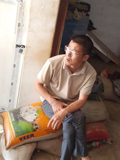 This image depicts a male individual seated indoors in Olomoda, Nigeria. The man, with short dark hair and wearing clear-framed glasses, a light tan polo shirt, and dark grey trousers, is positioned in the lower center, looking towards the upper right. His hands are resting on his lap. He is seated on a stack of sacks; the uppermost visible sack is orange, branded "VEER Indian Long Grain Parboiled Rice Product of India", and displays an image of cooked rice. To the immediate left of the man is a white door frame with a visible blue padlock secured to it. The background and right side of the frame reveal a dimly lit storage area containing numerous stacked sacks of varying colors (light-colored, blue, and red), along with a discarded plastic bottle visible on a concrete floor. The environment suggests a storeroom or a retail space for bulk goods.
